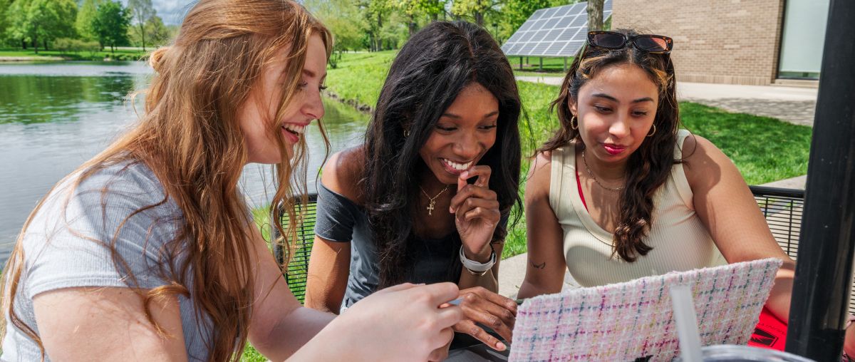 Three female students looking at a laptop outside in front of lake huntoon