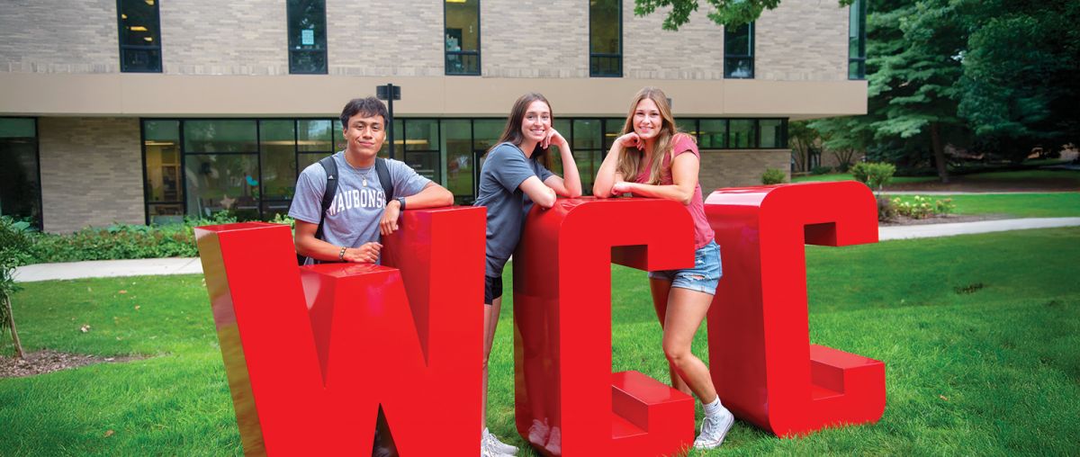 Three students standing with large WCC letters in front of Student Center in summer 2024