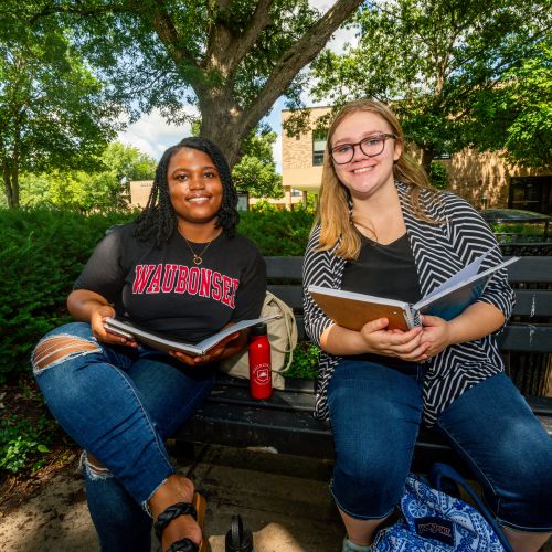 Two female students sitting outside on a bench with books open