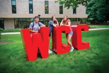 Three students standing with large WCC letters in front of Student Center in summer 2024